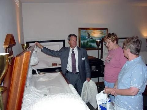 A man in a suit shows two women an open casket in a funeral home.