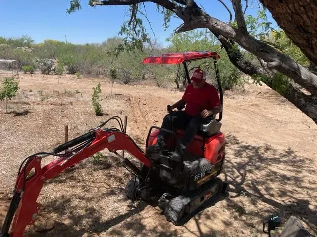 A man is driving a small red excavator in a field.