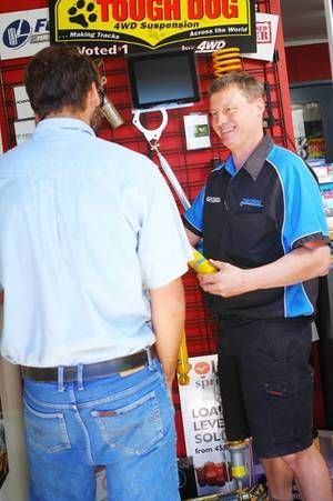 A staff member in a blue and black uniform shows a yellow component to a customer in a store with auto suspension parts.