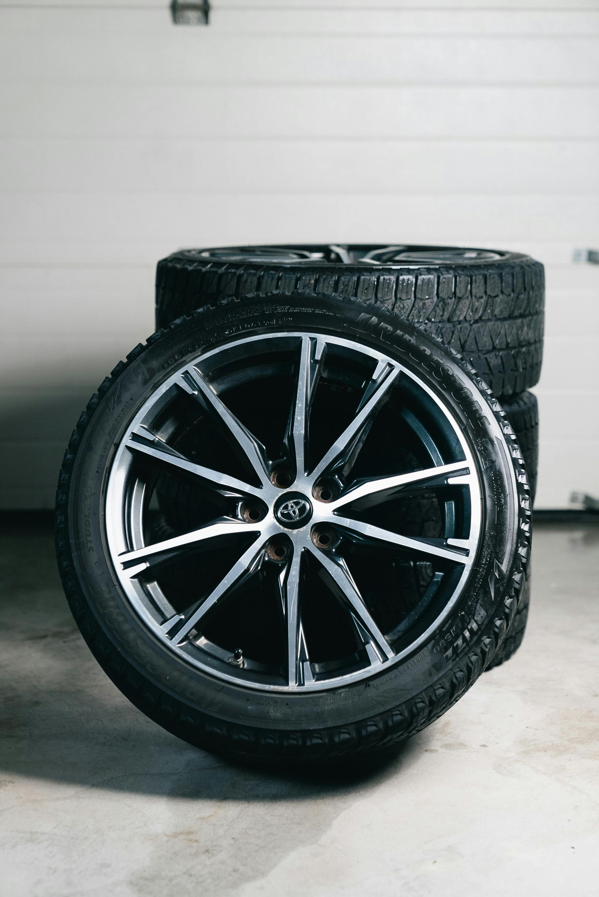 A front-facing car wheel with a sleek silver and black multi-spoke rim, set against another stacked tire in a garage.