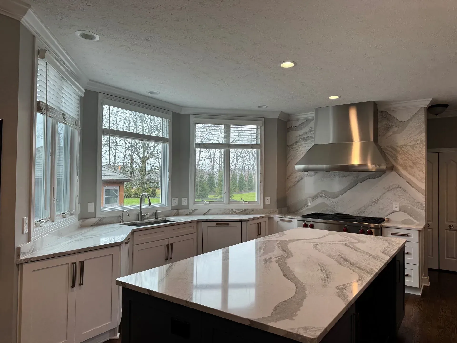 A modern kitchen with a white marble countertop island and range hood, with a windowed backsplash.