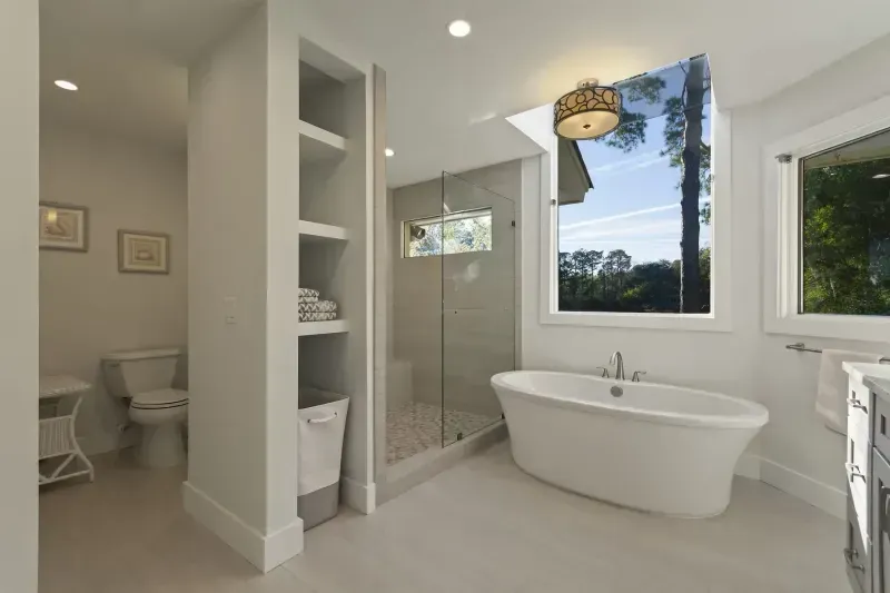 Bathroom with a white freestanding tub, shower, toilet, and built-in shelving. Large window with outdoor view.