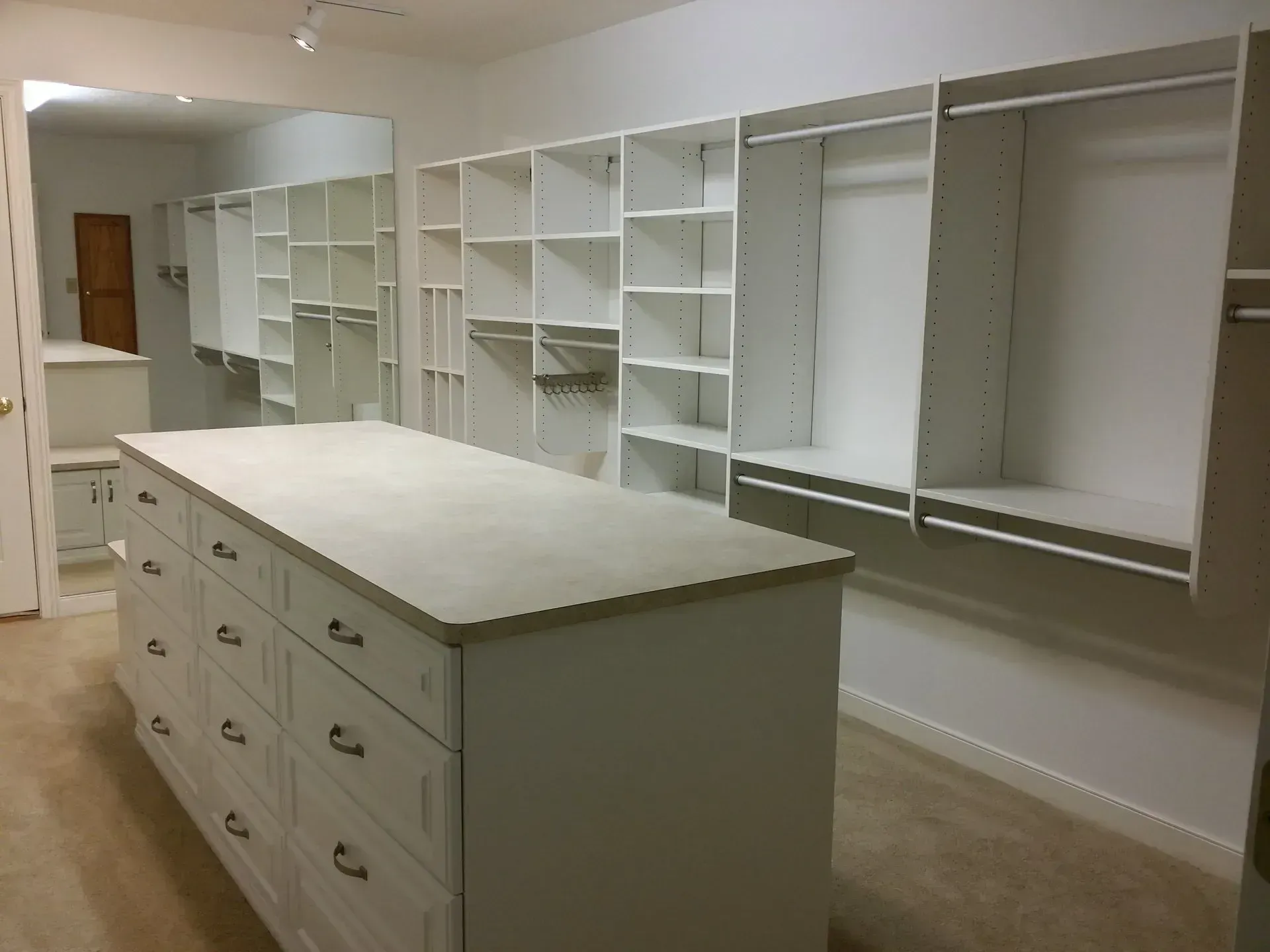 Walk-in closet with white shelves, drawers, and an island. Beige carpet and a large mirror.