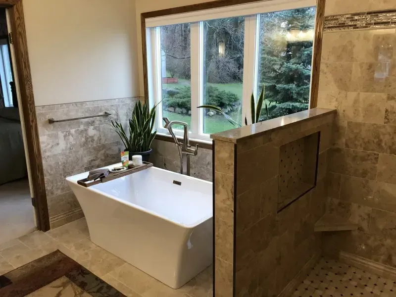Modern bathroom with a white tub, natural stone tile, and a large window overlooking a green yard.