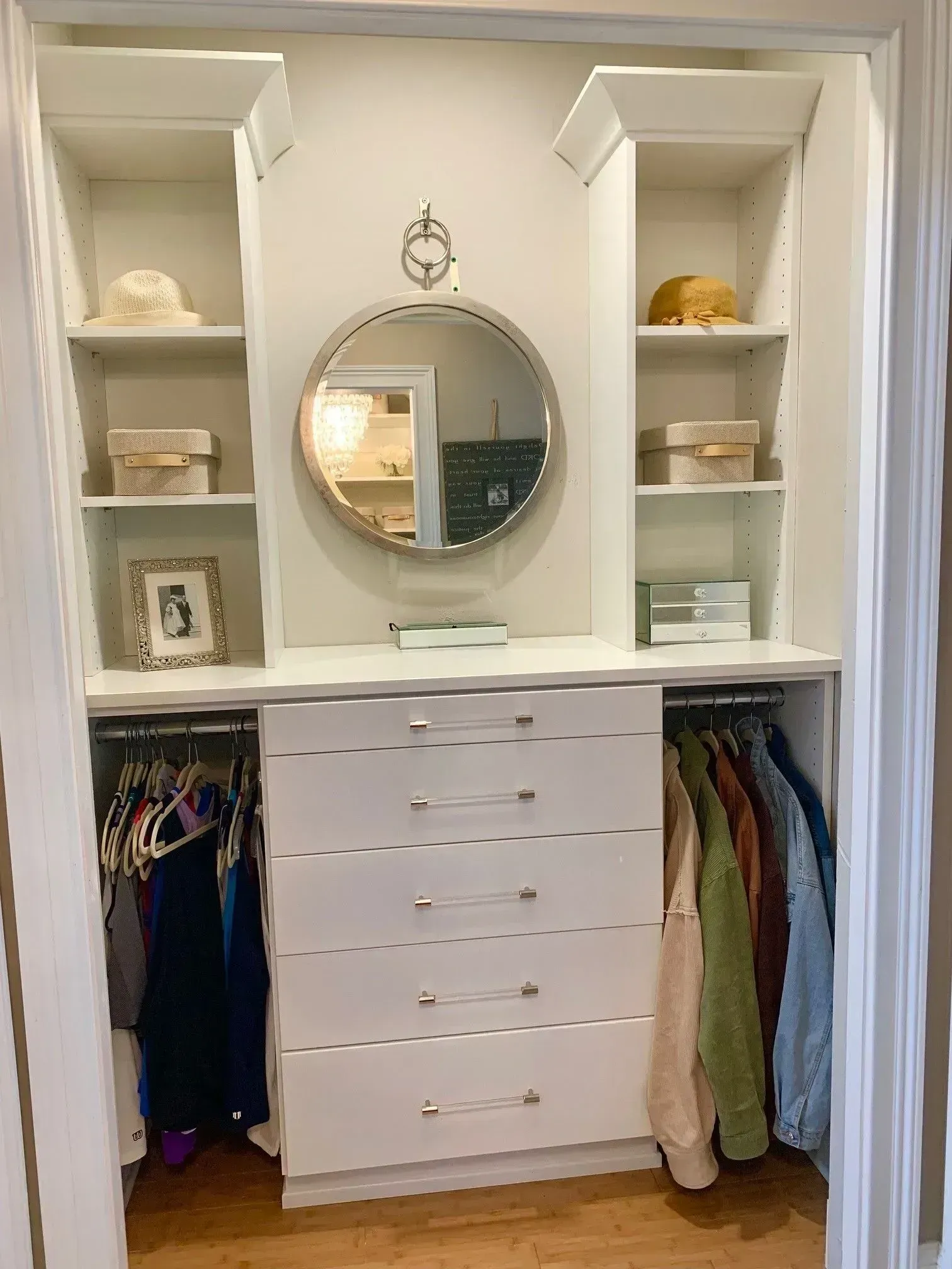 White closet interior with built-in shelves, drawers, and hanging rods. Round mirror centered with decor.