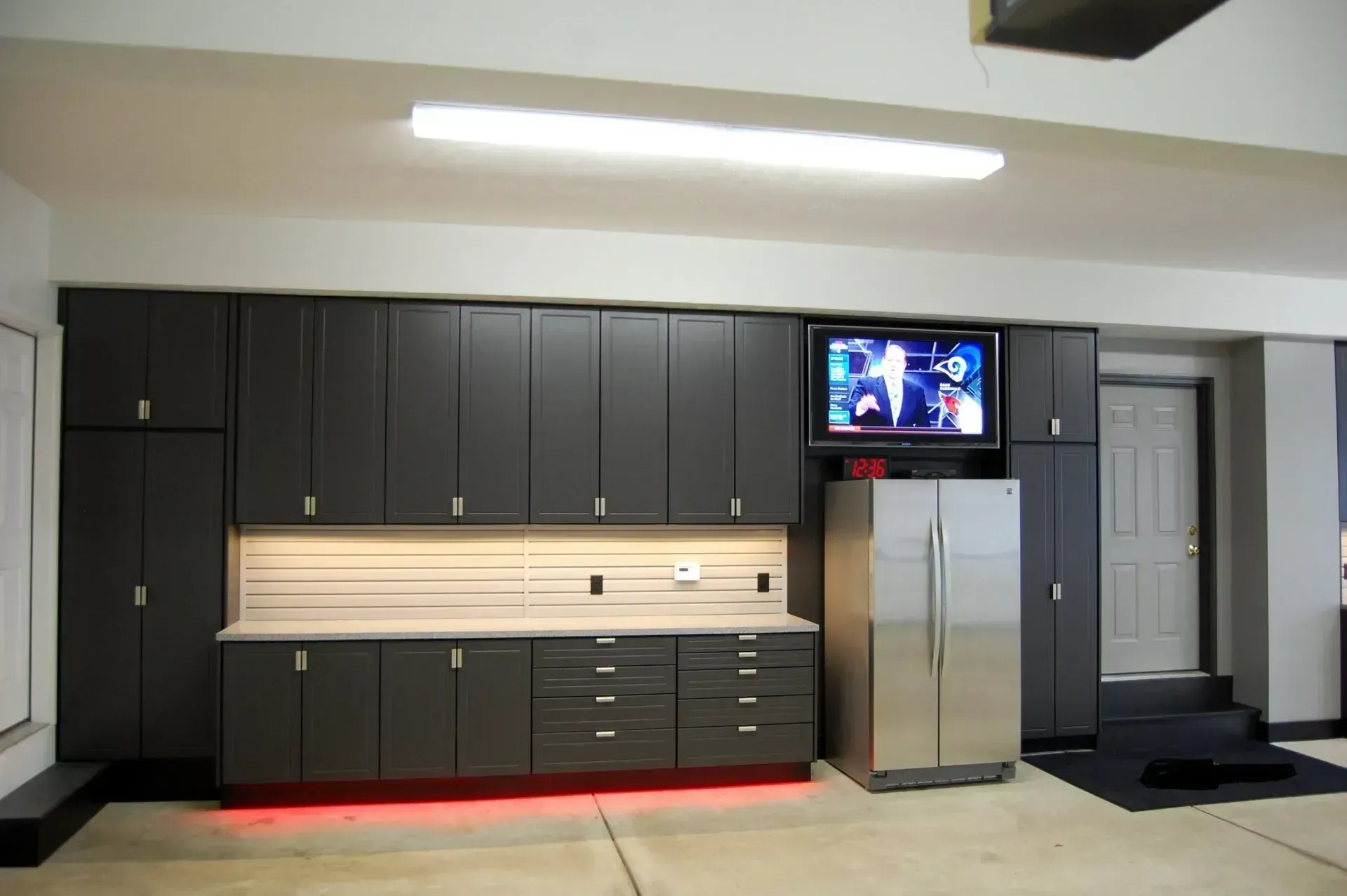 Garage with dark gray cabinets, refrigerator, TV, and work surface with red under-cabinet lighting.