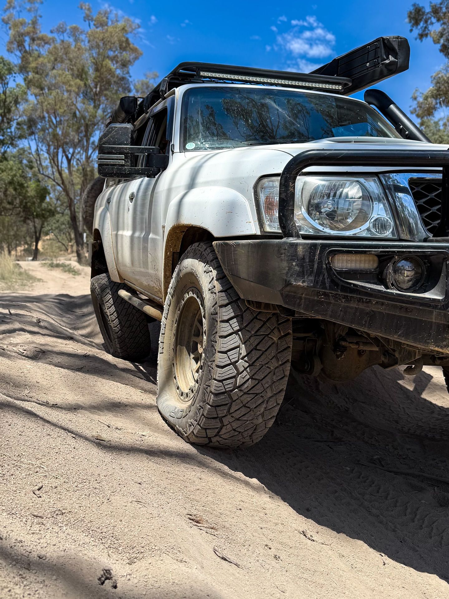 White off-road vehicle with a flat tyre on a dirt road, trees in the background under a blue sky. — Atlas 4X4 Touring & Mechanical in Tweed Heads, NSW