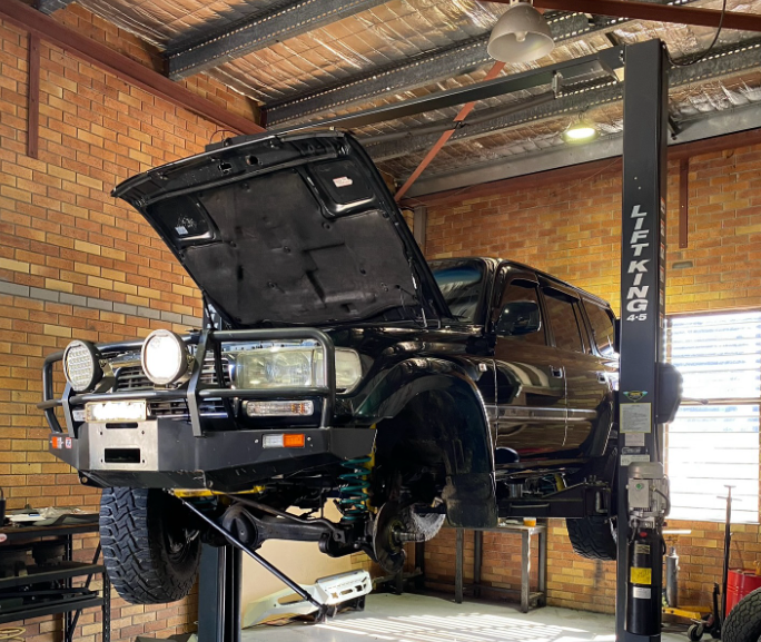 Black SUV on a lift with the hood open in a garage, lights and brick wall visible. — Atlas 4X4 Touring & Mechanical in Tweed Heads, NSW