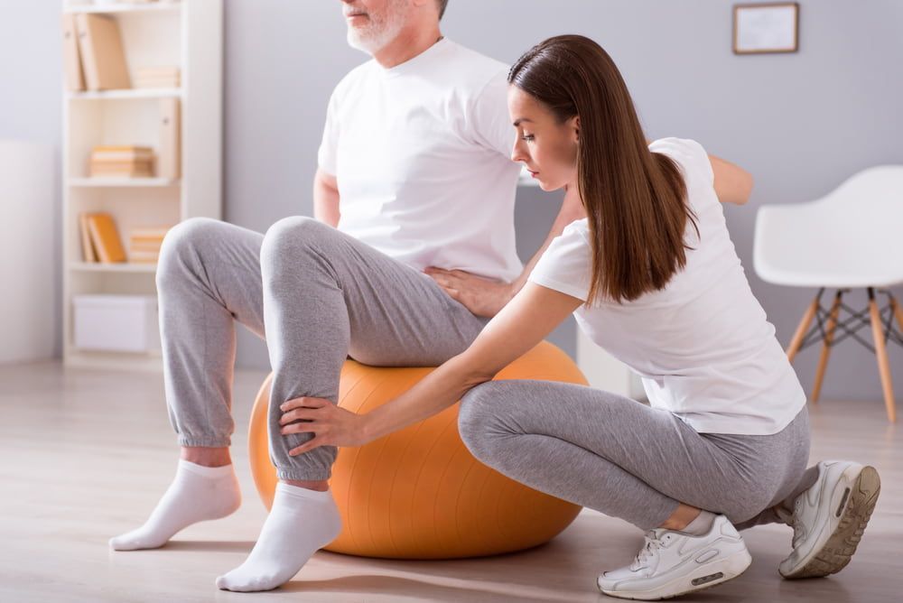 A Woman Is Helping A Man With A Exercise Ball — Southern Districts Physiotherapy & Sports Injuries Clinic In Albion Park, NSW