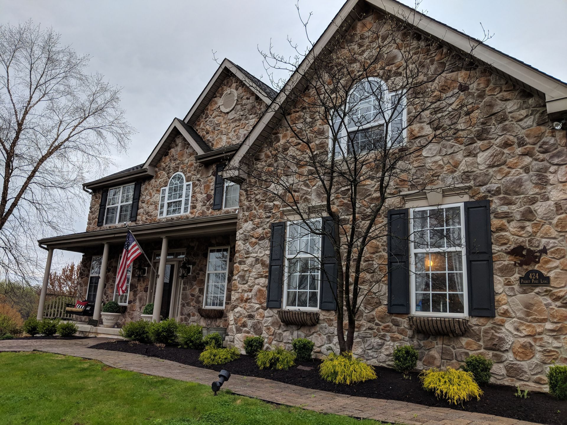 A large stone house with white windows black shutters and a flag on the front porch.
