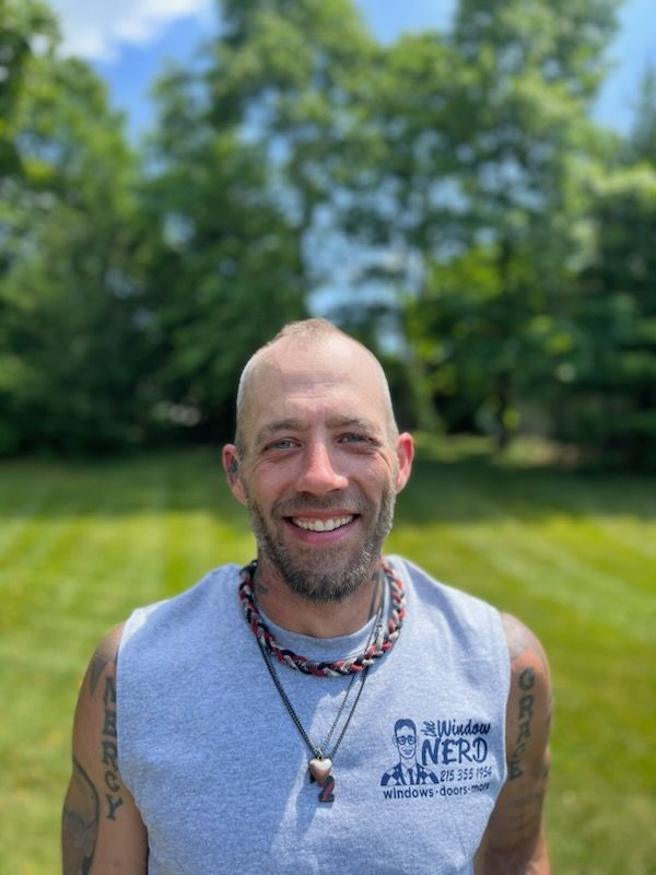 A man wearing a gray tank top and a necklace is smiling in a field in Doylestown, PA.