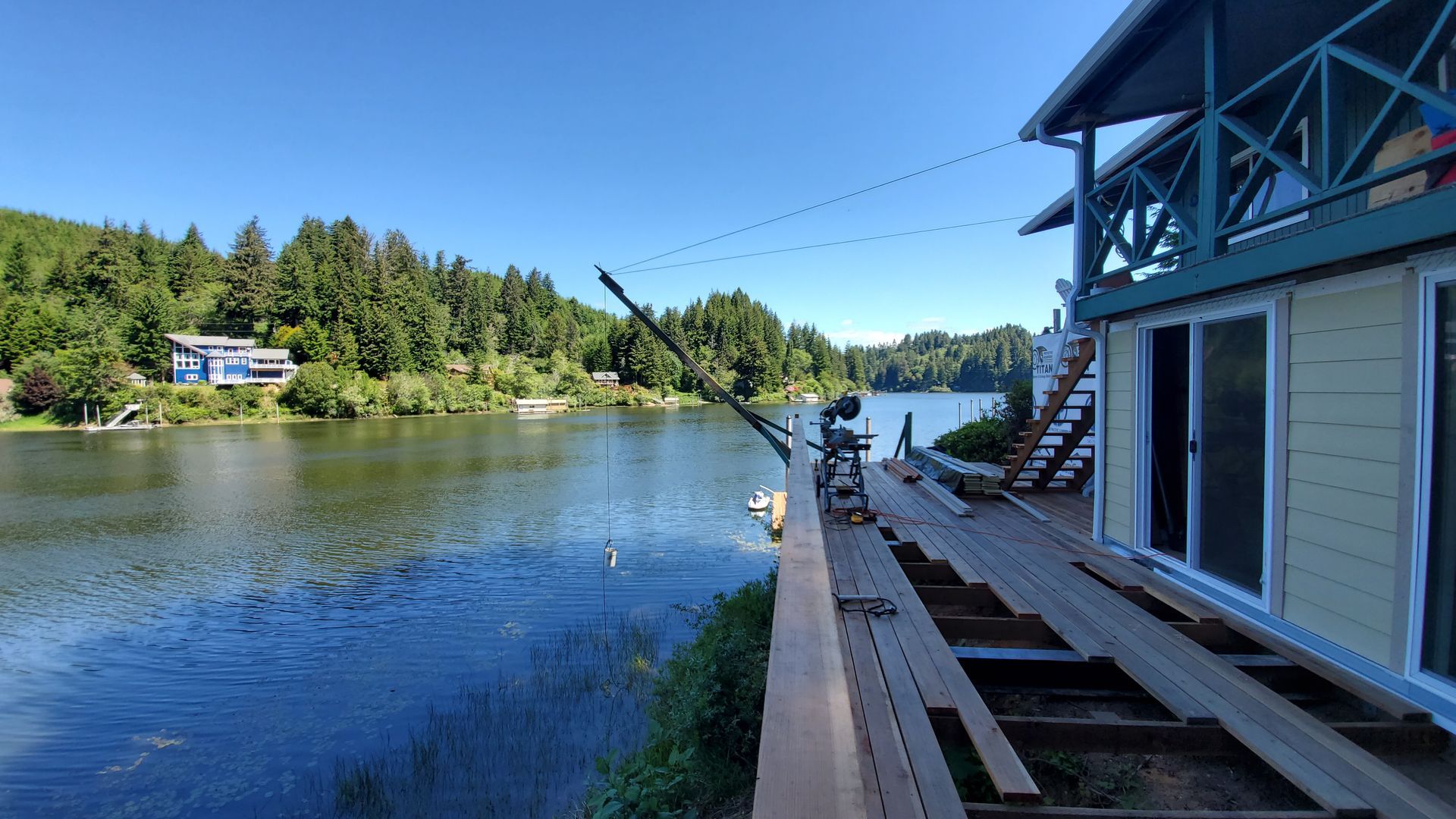 Lakeside view with a house and deck. Blue water, trees, and a sunny sky.
