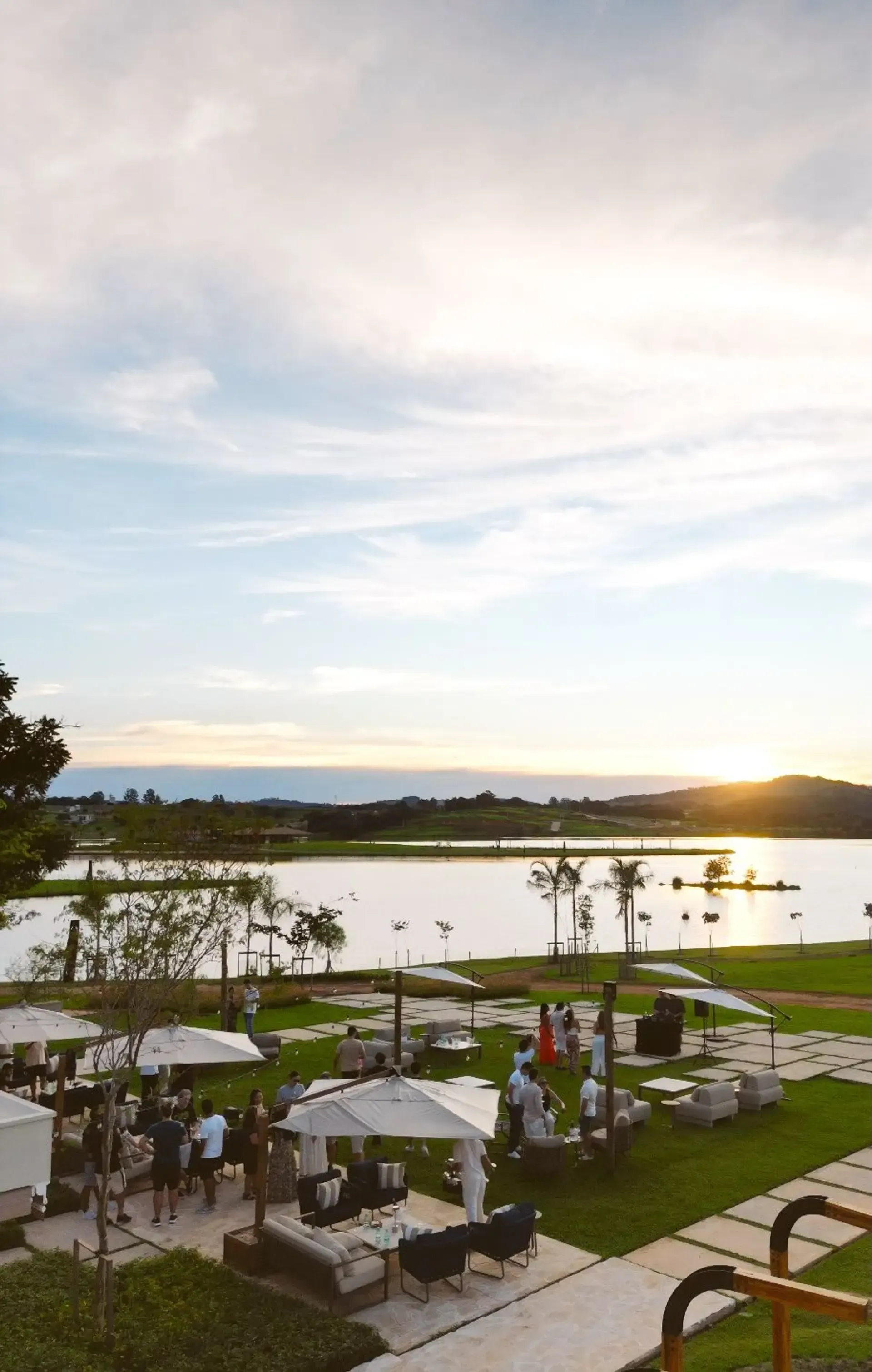 Pôr do sol sobre um lago com pessoas socializando em uma área gramada com copas brancas.