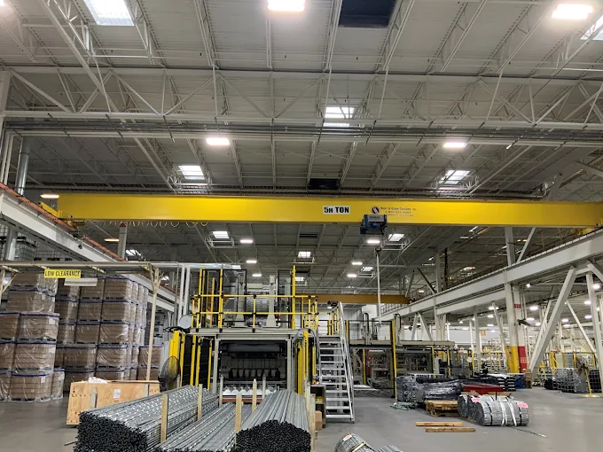 Yellow overhead crane in a factory with machinery and stacks of metal materials.