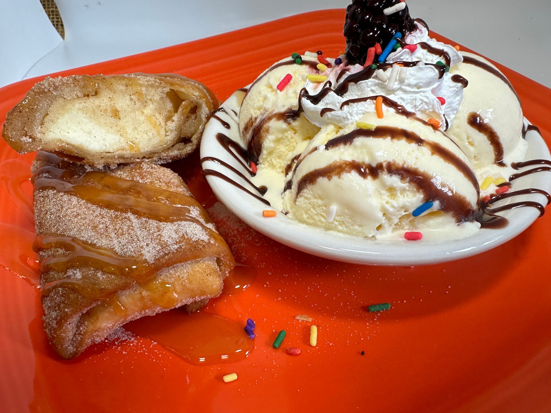 Fried pastry and sundae with vanilla ice cream, chocolate syrup, whipped cream, sprinkles, and a cherry on an orange plate.