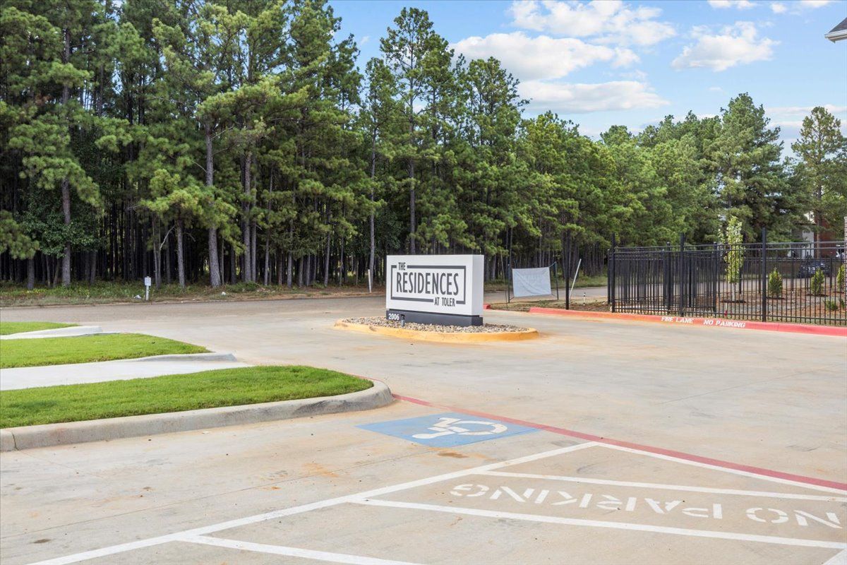 Entrance to a residential complex with sign, wheelchair accessible parking, and trees.