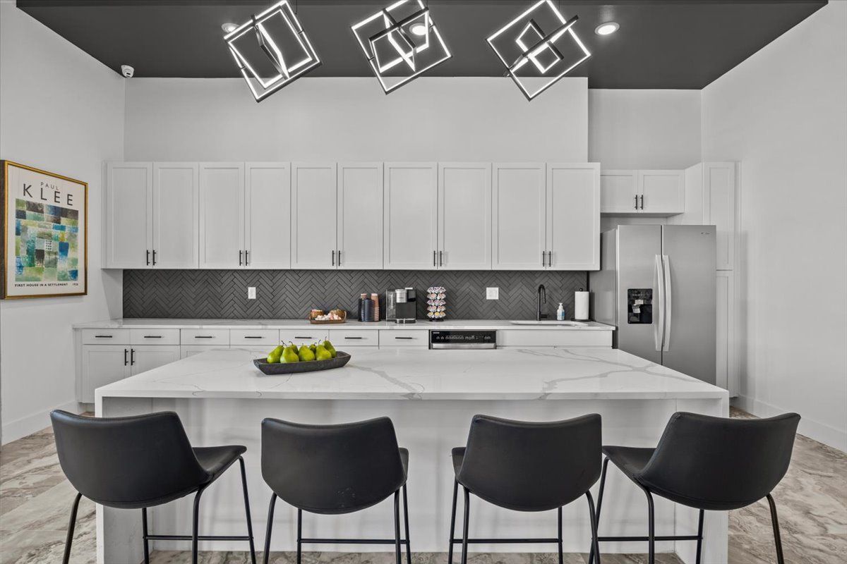 Modern white kitchen with island, black chairs, cabinets, stainless steel appliances, and decorative lighting.