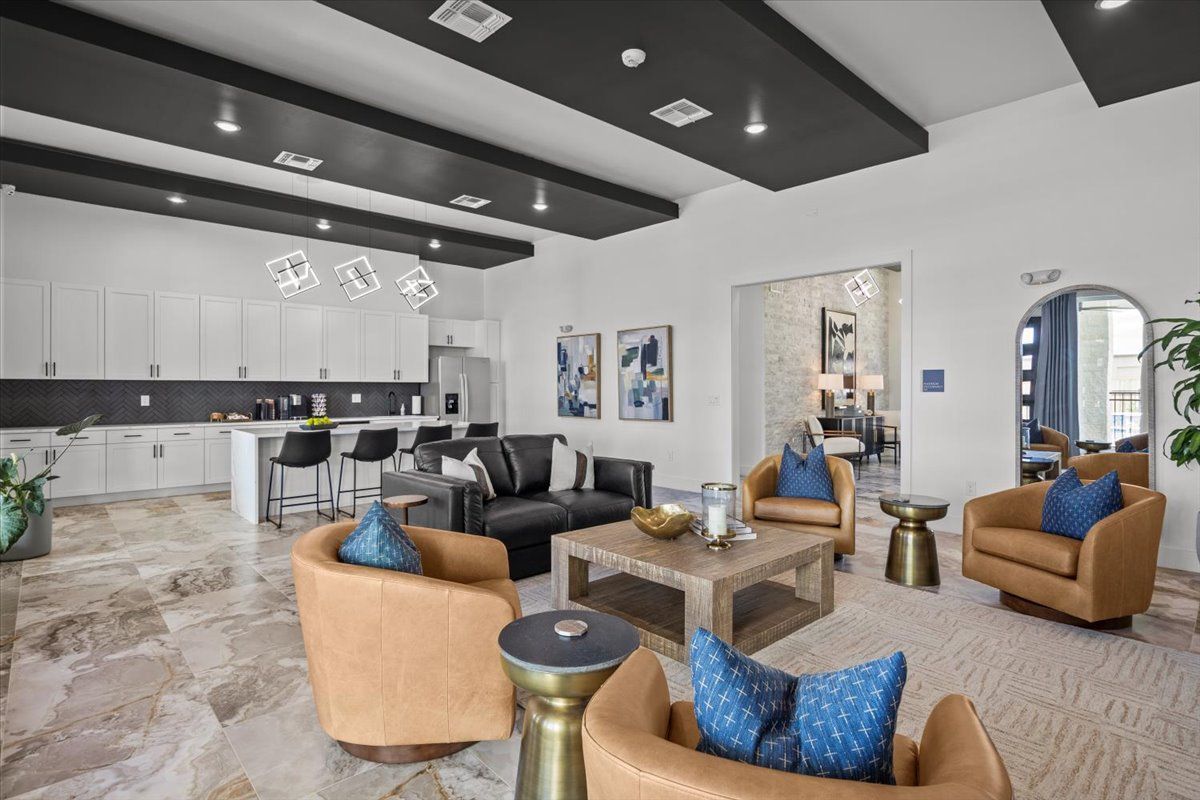 Living room with kitchen. Tan leather chairs with blue pillows, dark couch, and white cabinets.