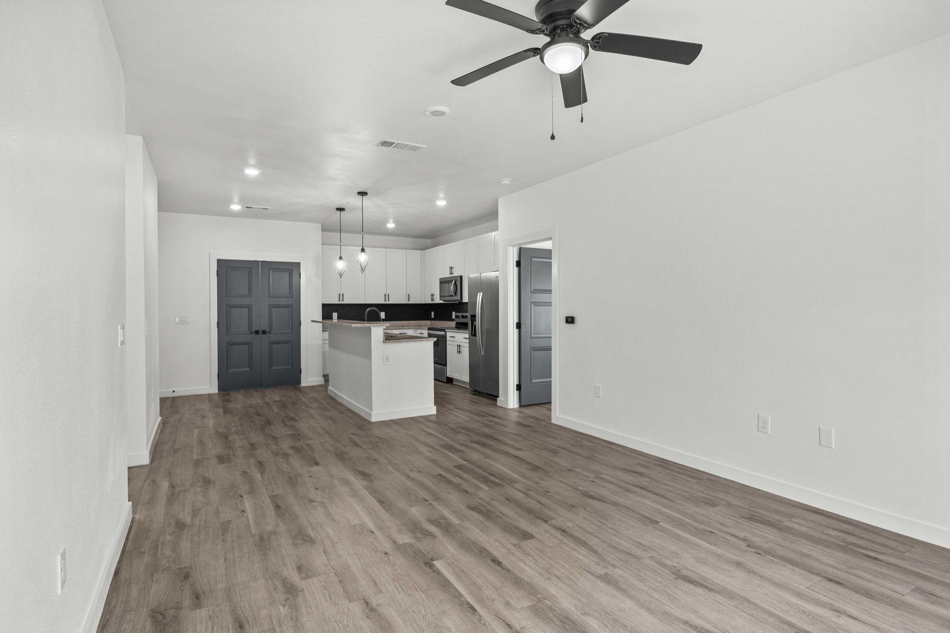 Spacious, empty living area with open kitchen; grey wood-look floors, white walls and cabinets, and dark grey doors.