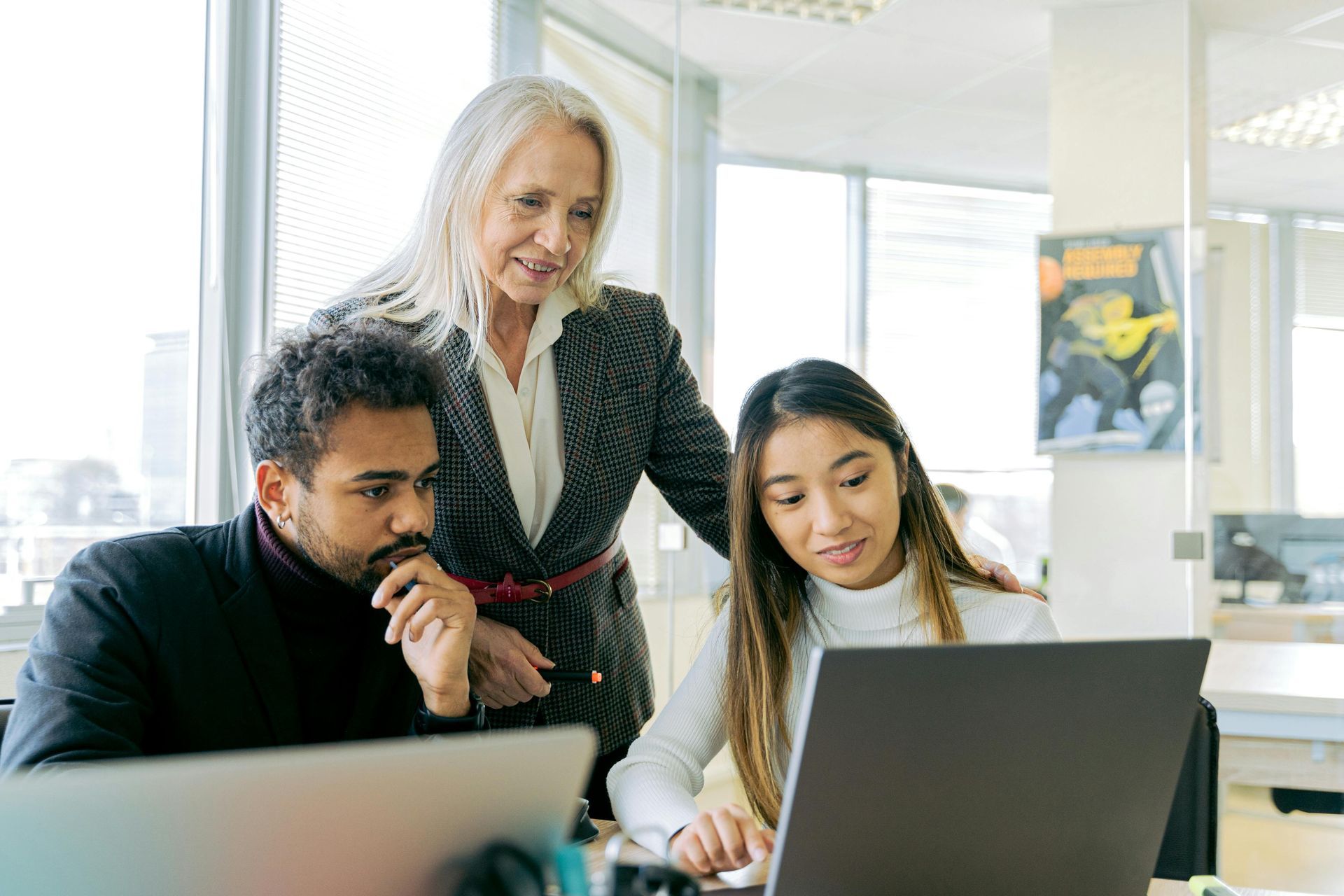 Three people looking at laptop screens in an office setting. One woman leans over the shoulder of a younger woman, while a man looks on.
