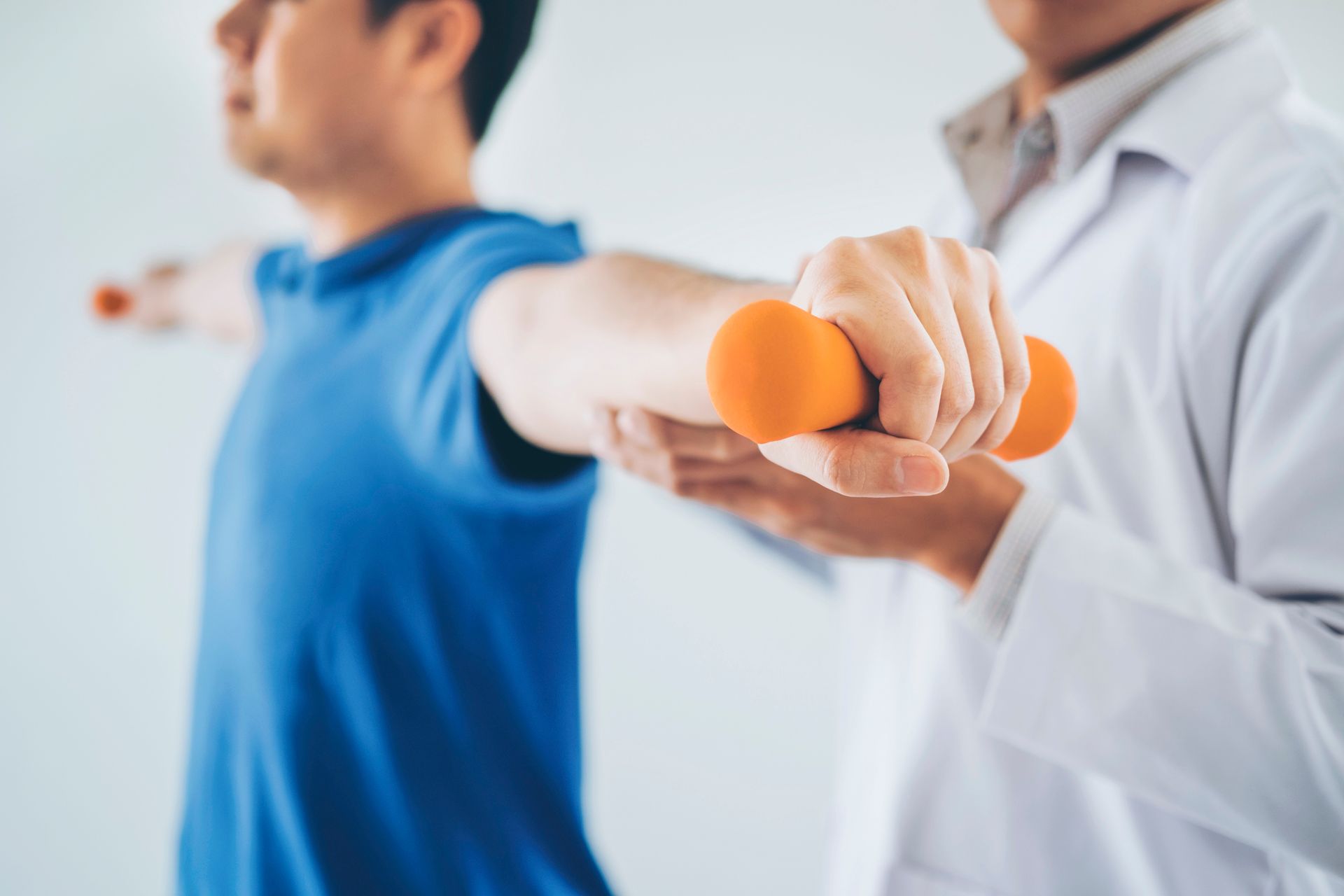 Man in blue shirt doing arm exercise with dumbbell, guided by a person in a white coat.