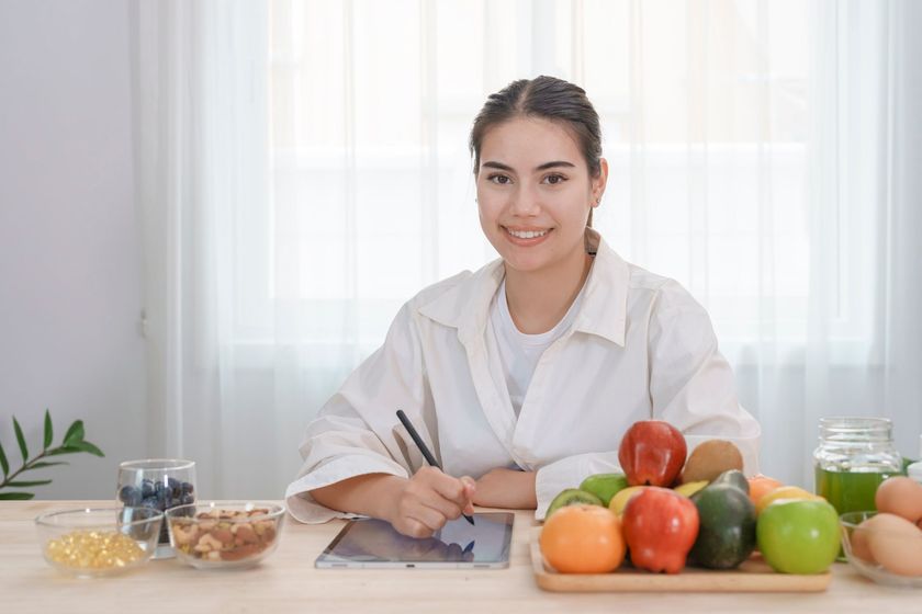Woman smiling, drawing on tablet, surrounded by fruit, nuts, and supplements on a table.