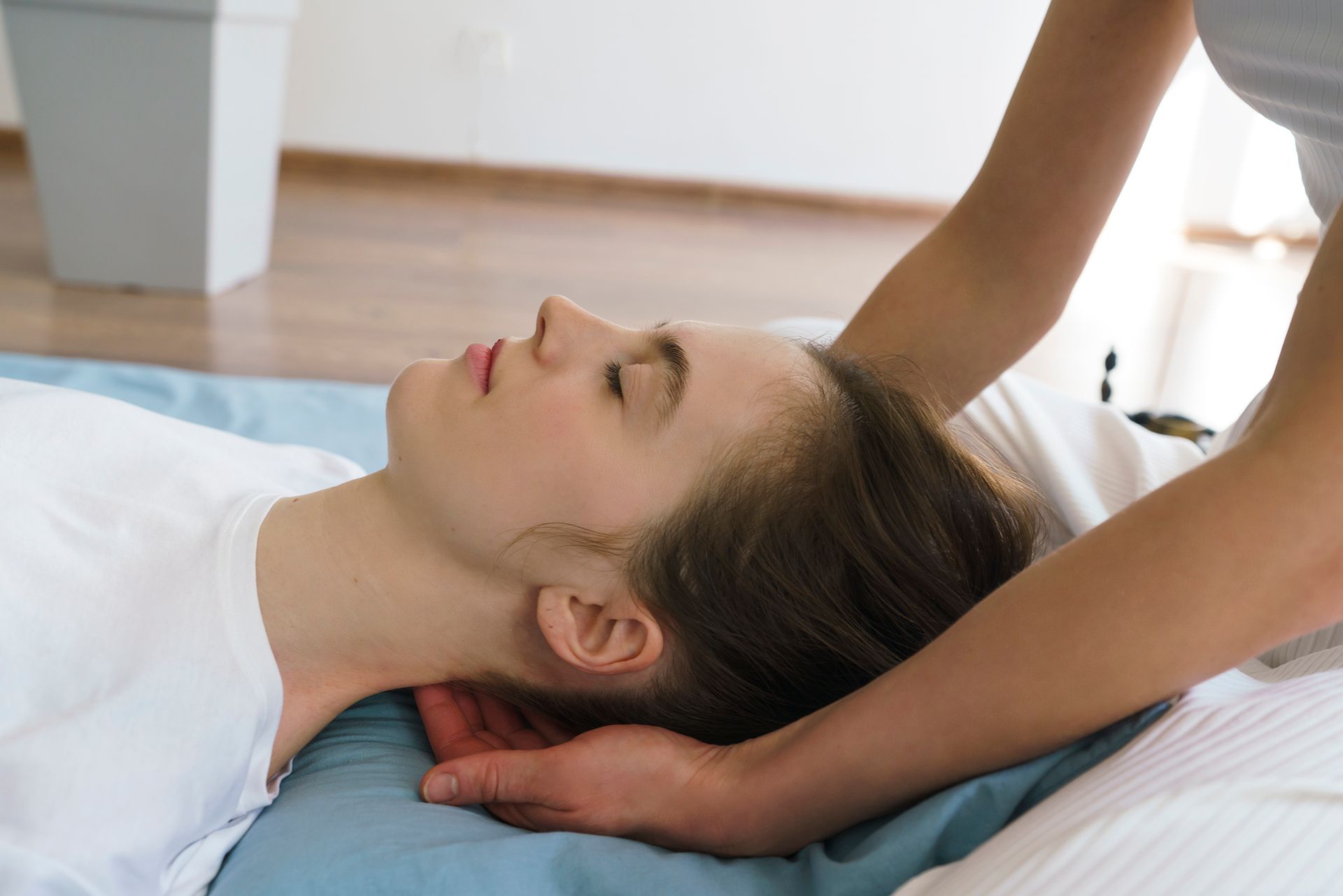 Person receiving a neck massage, eyes closed, on a blue mat. Another person's hands cradle their head. Person receiving a neck massage, eyes closed, on a blue mat. Another person's hands cradle their head.