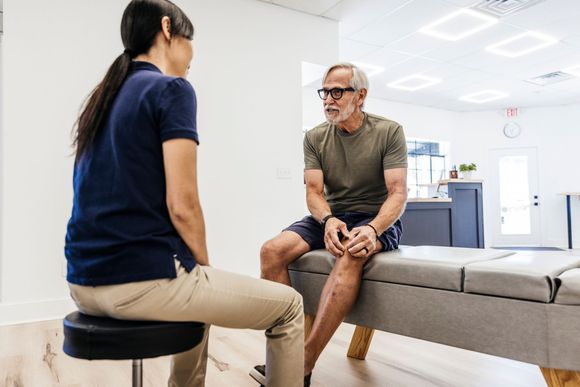 A therapist in a blue shirt talking with a patient seated on a therapy table, in a clinic room.