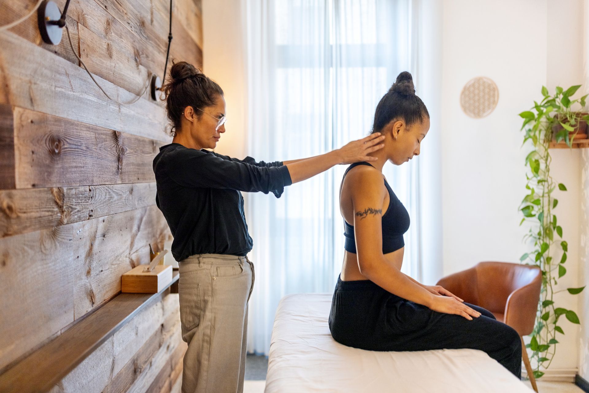 Woman performing a neck massage on another woman seated on a massage table. Wooden wall backdrop. Woman performing a neck massage on another woman seated on a massage table. Wooden wall backdrop.