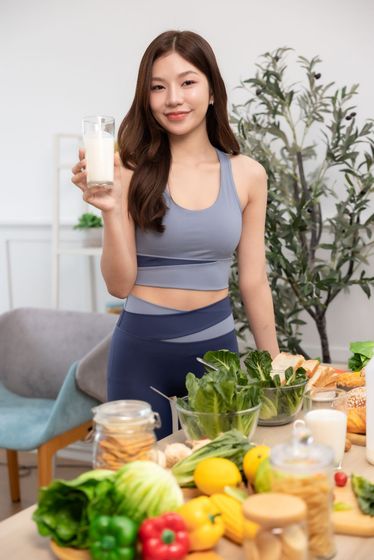 Woman in workout clothes holding a glass of liquid, surrounded by vegetables and healthy food.