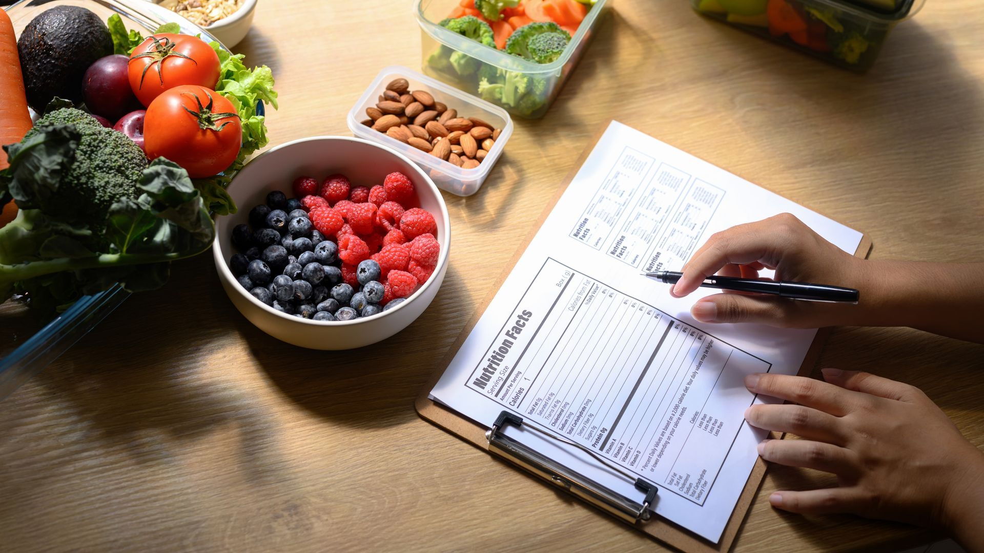Person writing on a nutrition label with fresh fruits, vegetables, and nuts.