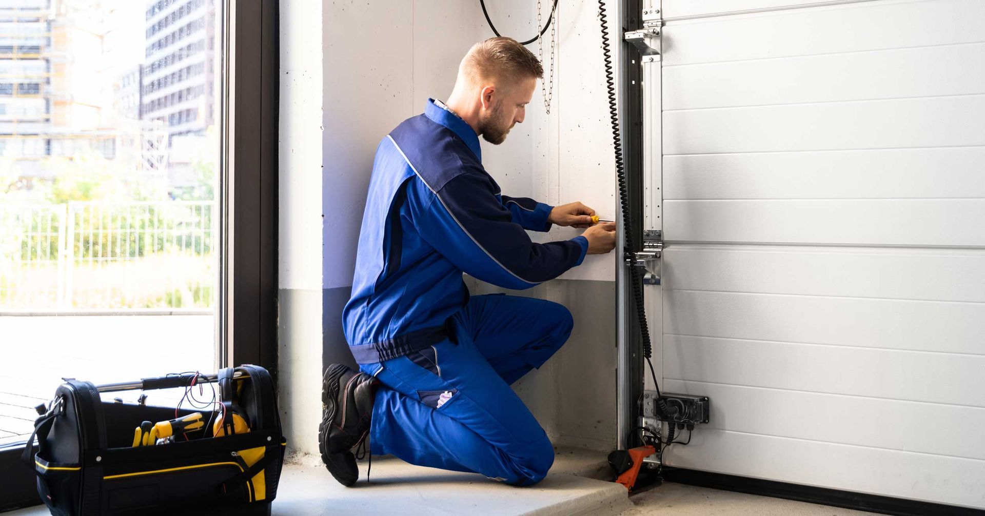A technician in a blue uniform kneels to repair the bottom section of a garage door track.