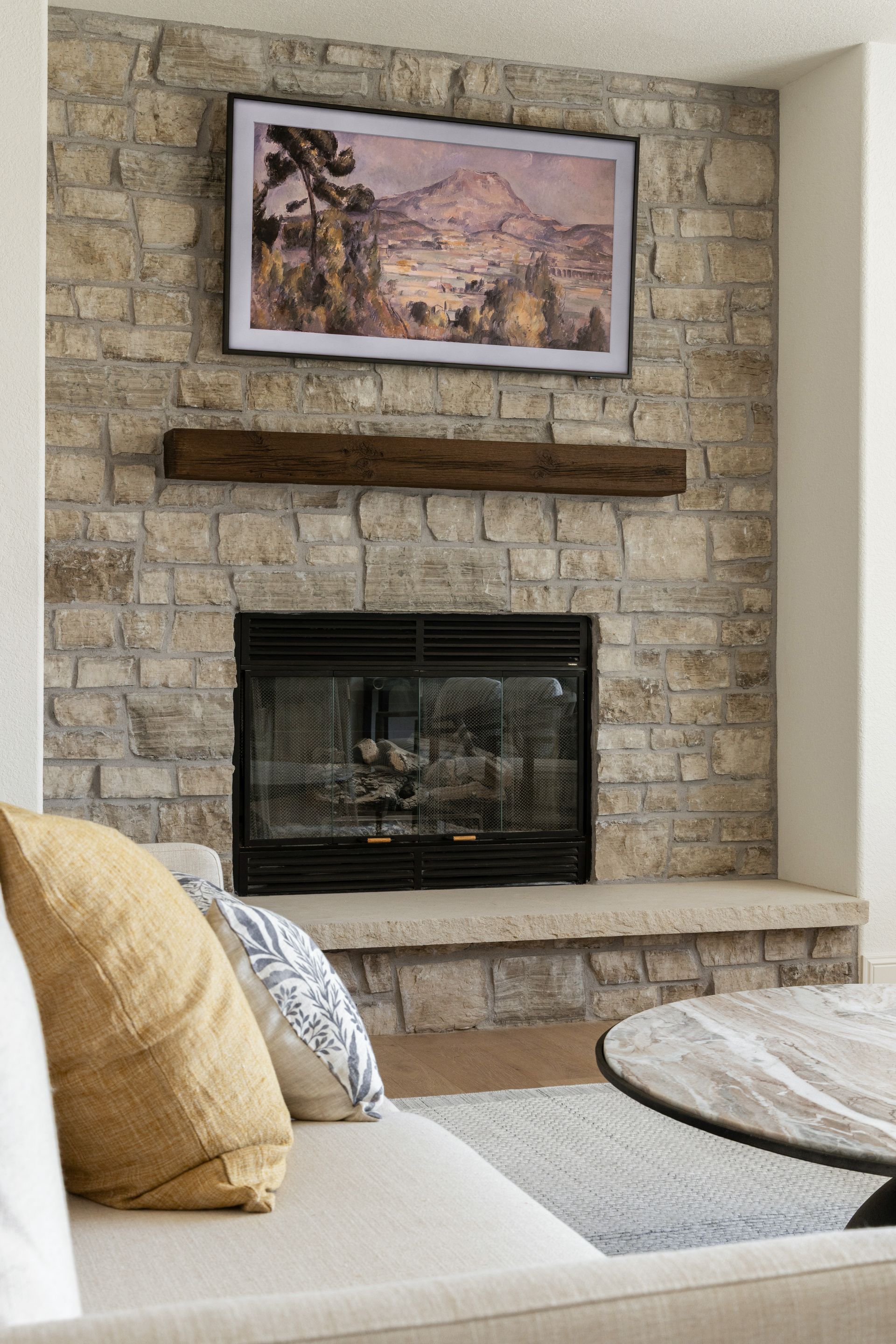 Fireplace with stone surround, TV displaying art above wooden mantel. Beige couch with pillows in foreground.