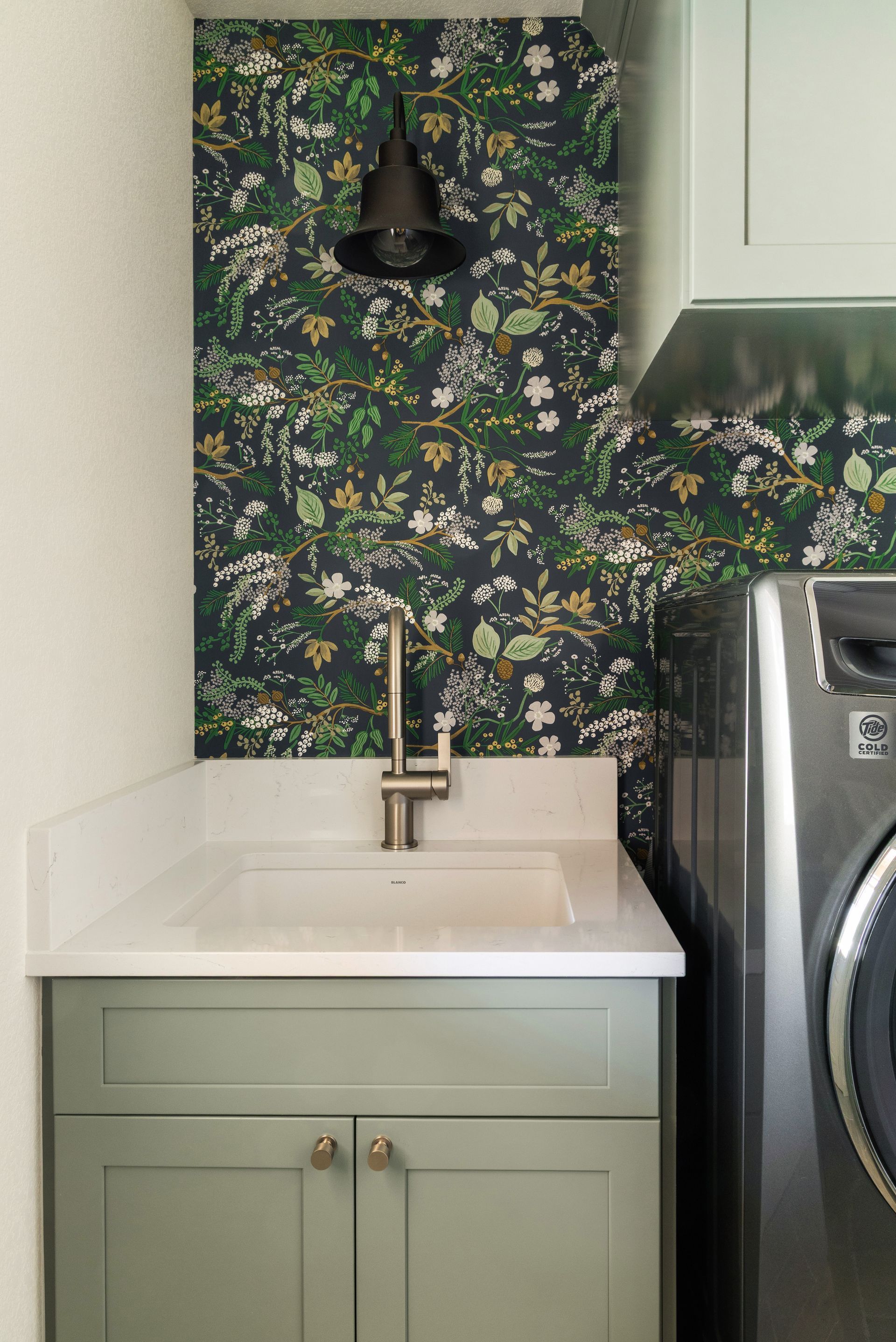 Laundry room with sage green cabinets, floral wallpaper, and silver faucet.
