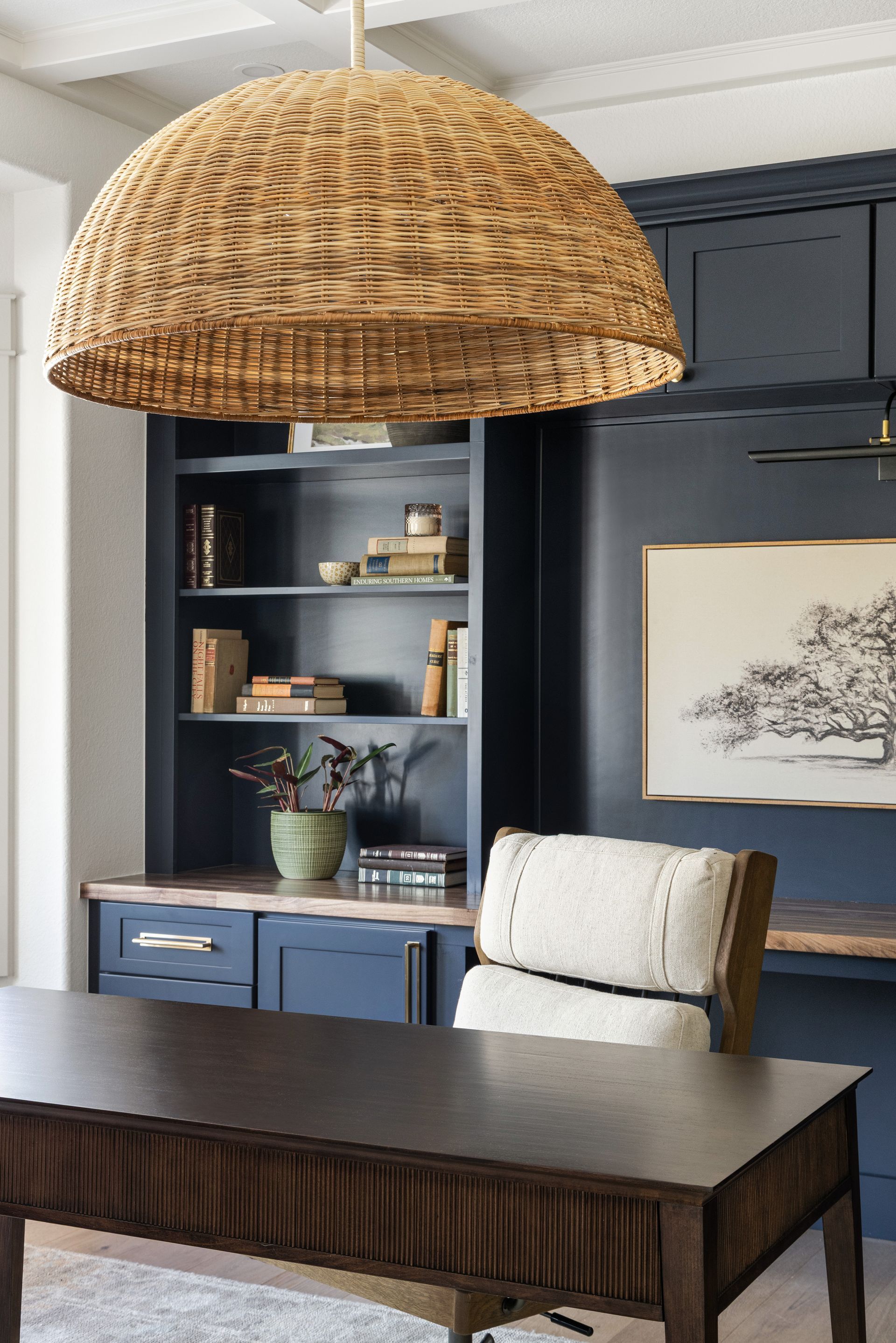 Home office with dark blue cabinets, wooden desk, woven pendant lamp, and chair.