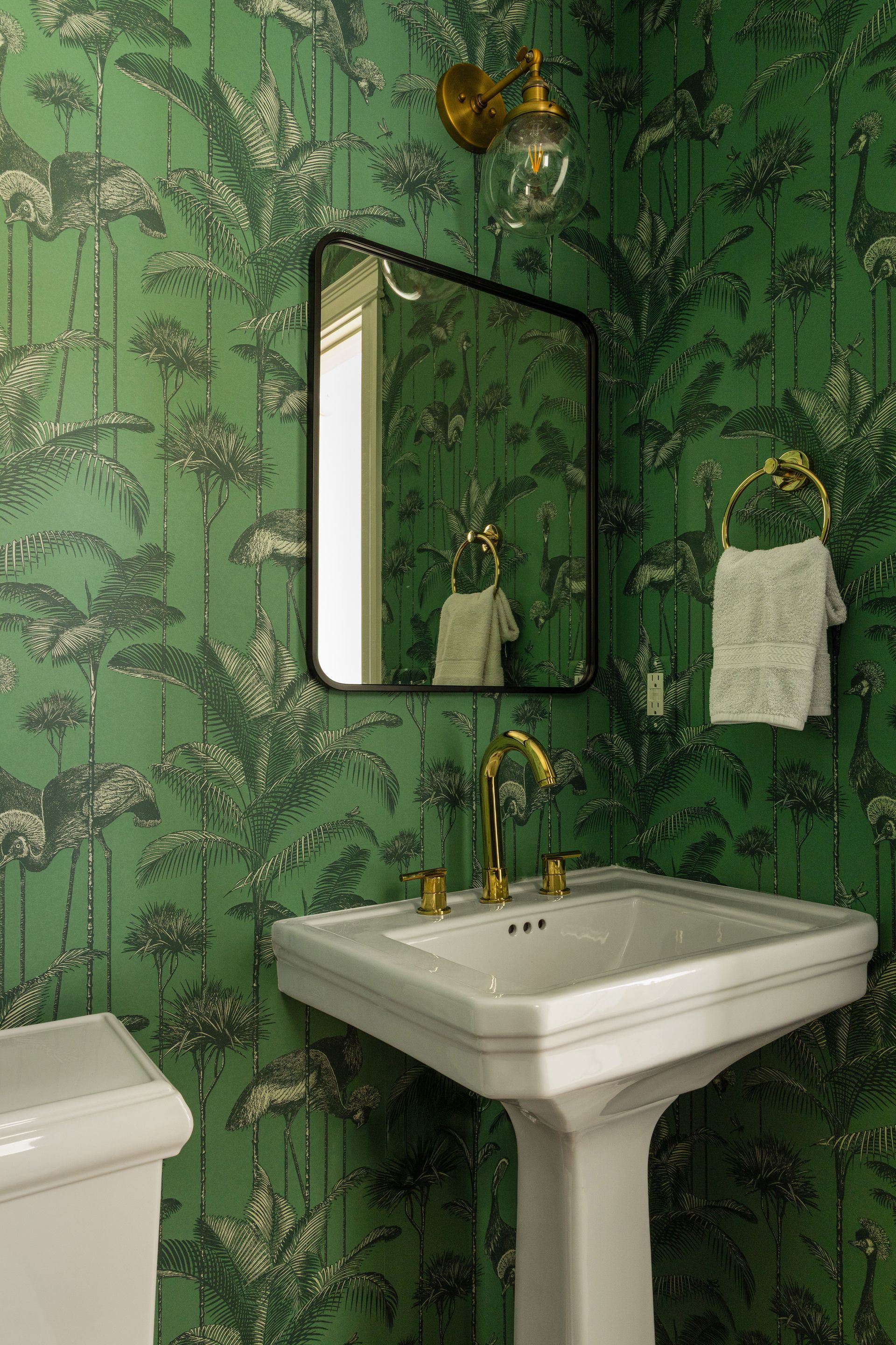 Green tropical wallpapered bathroom with white pedestal sink, black-framed mirror, and gold fixtures.