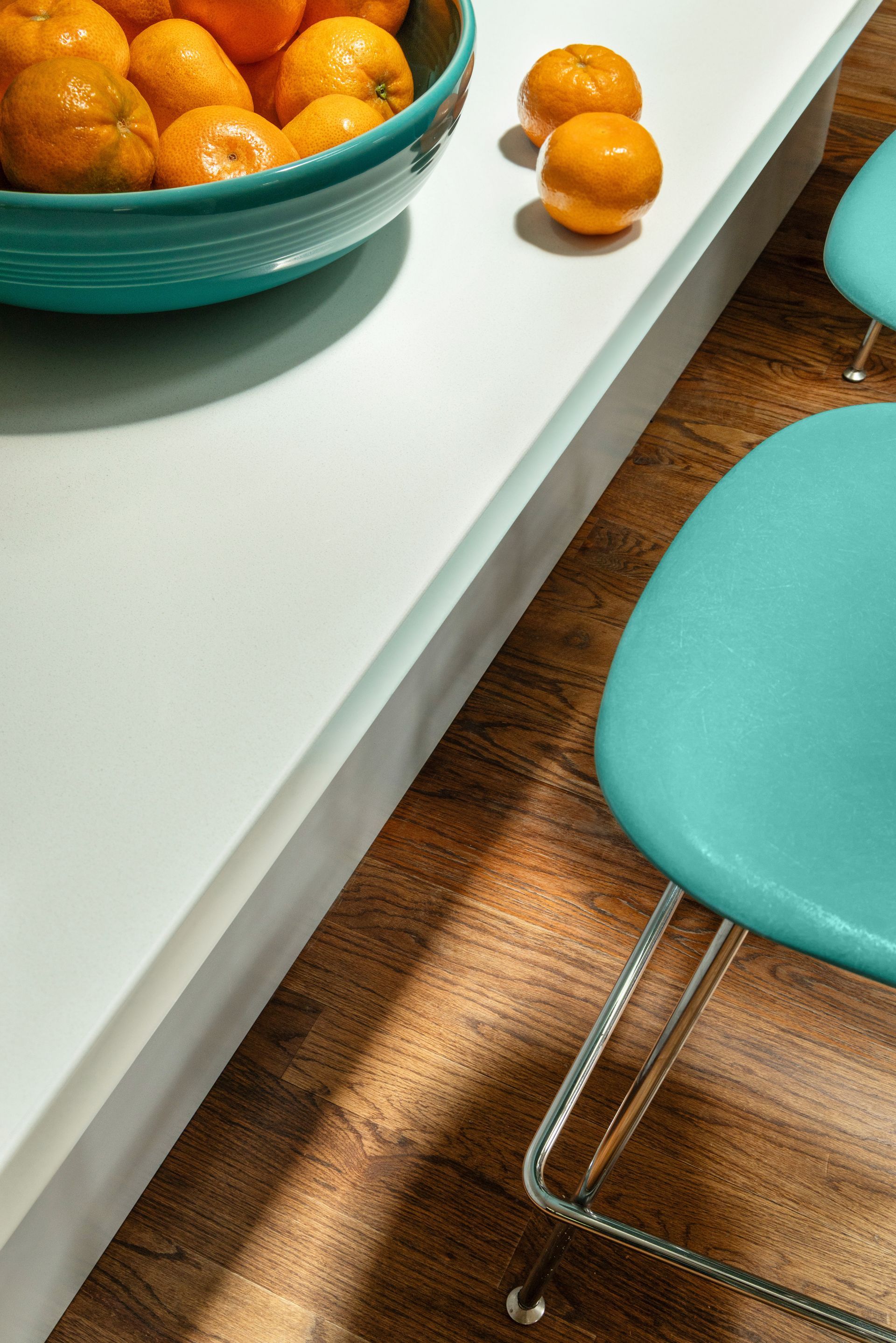 Close-up of a teal bowl of oranges on a white counter, teal chairs on a wood floor.