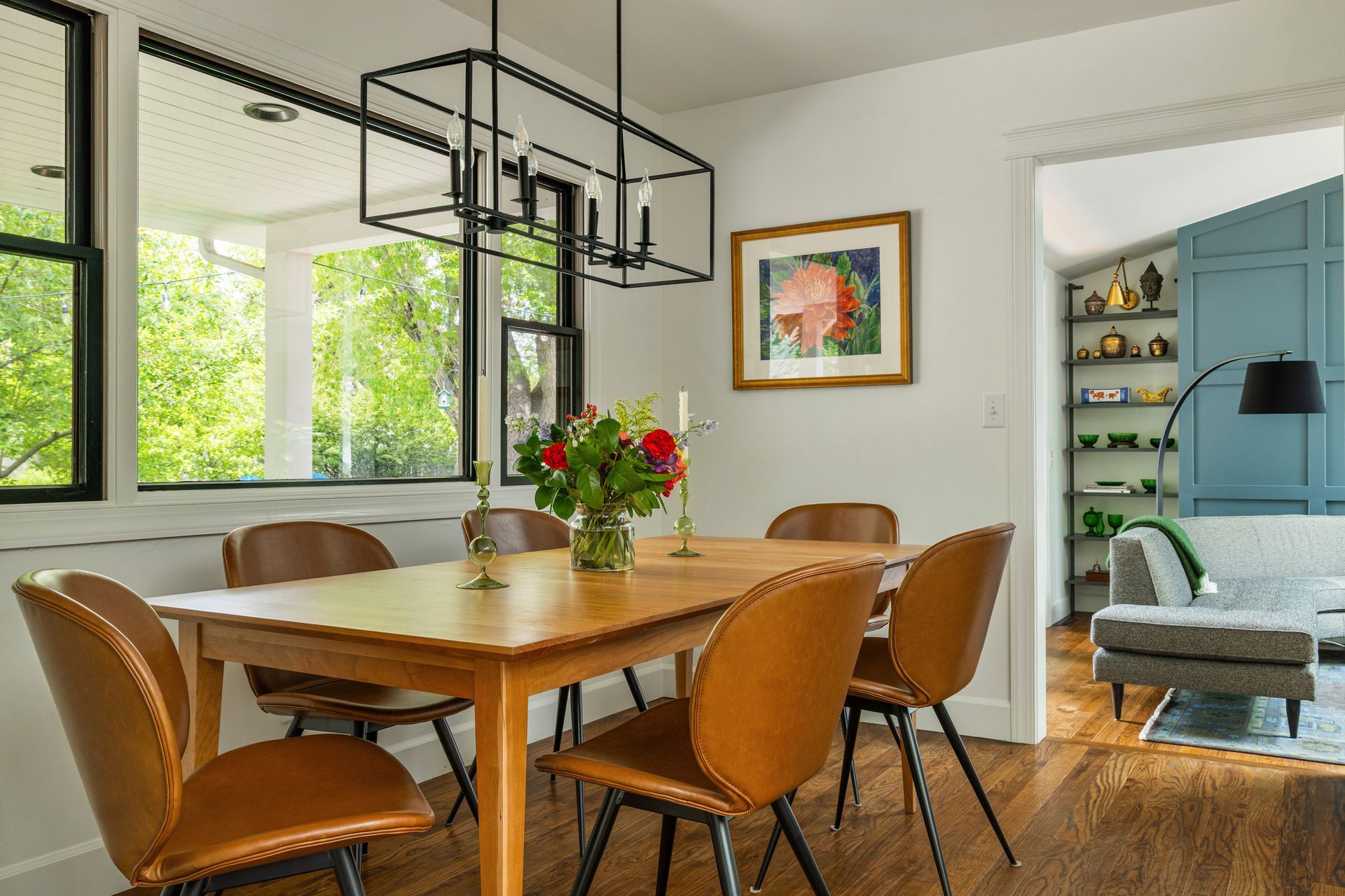 Dining room with wooden table and brown chairs, black chandelier, and artwork.  View into a living room.
