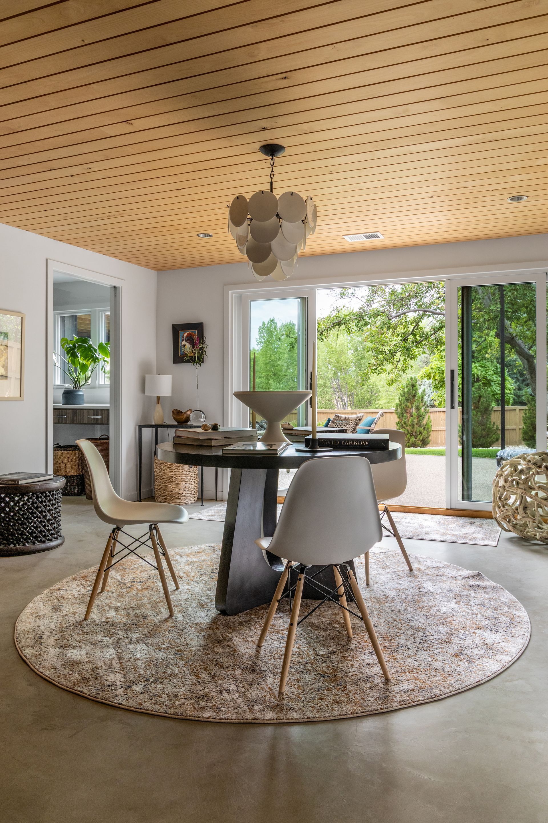 Round dining room with wooden ceiling, round rug, modern chairs, and view of a garden through sliding doors.