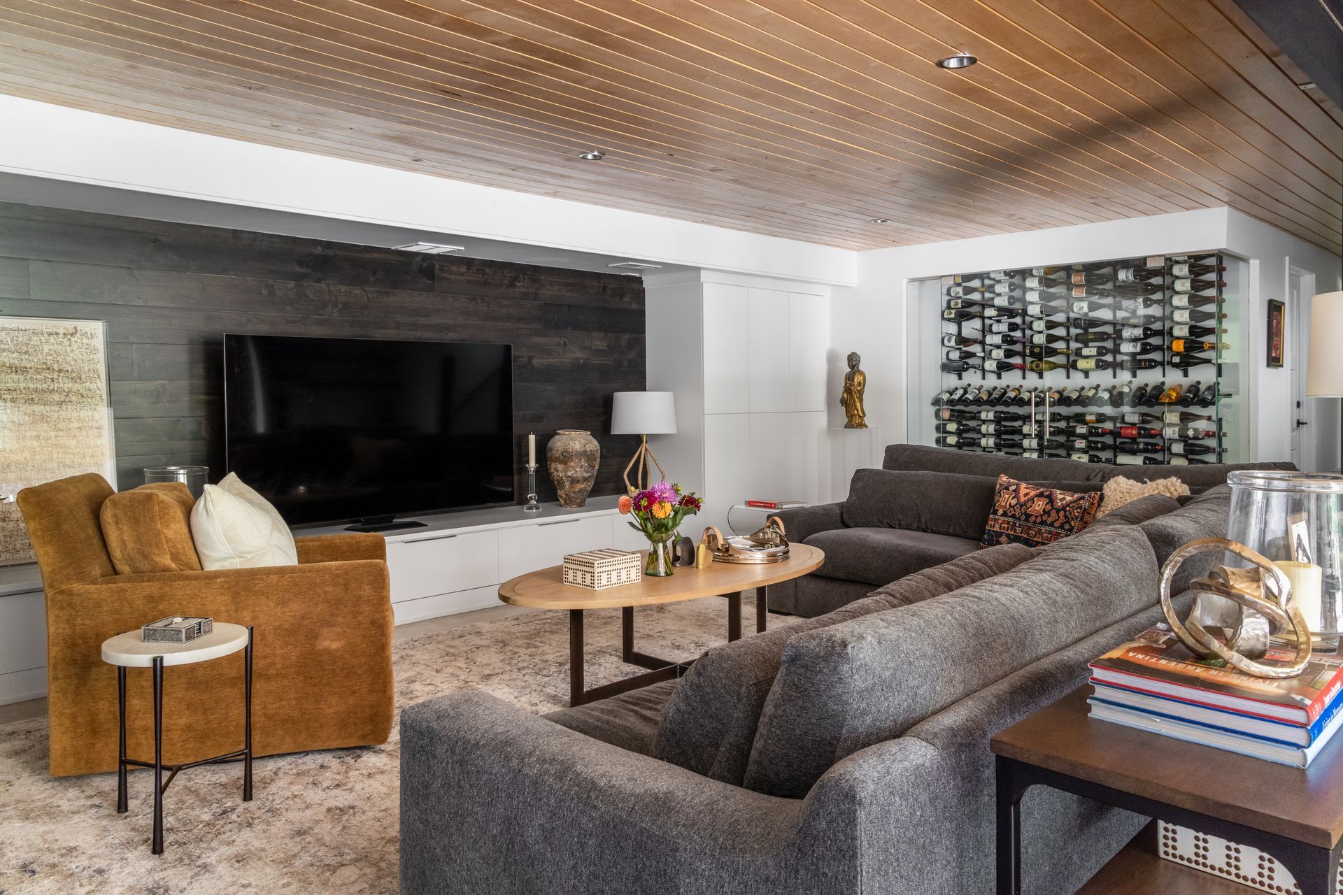 Cozy living room with gray sectional sofa, TV, wine rack, and wood ceiling.