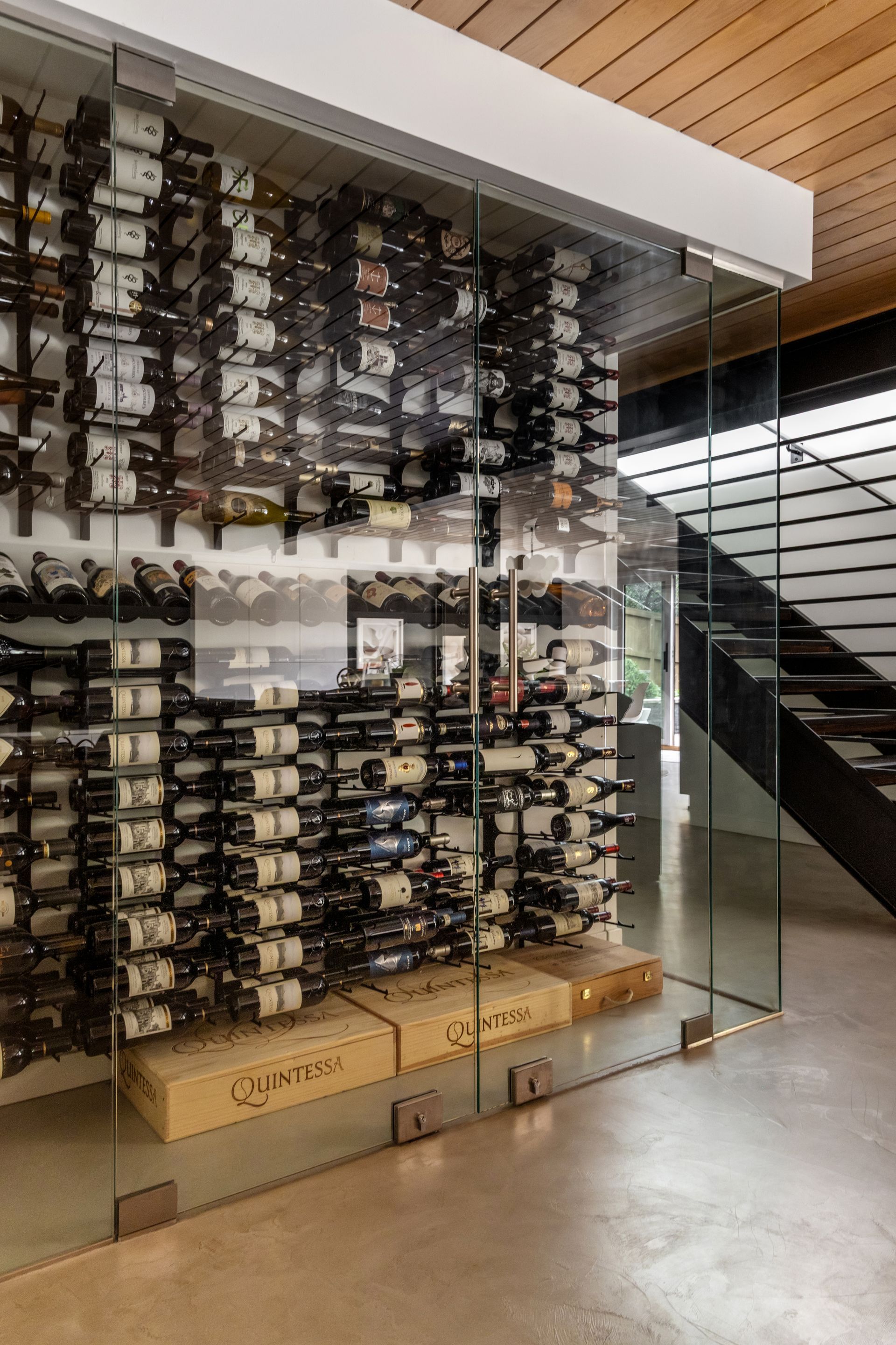Wine cellar with glass walls, filled with bottles, next to a staircase.
