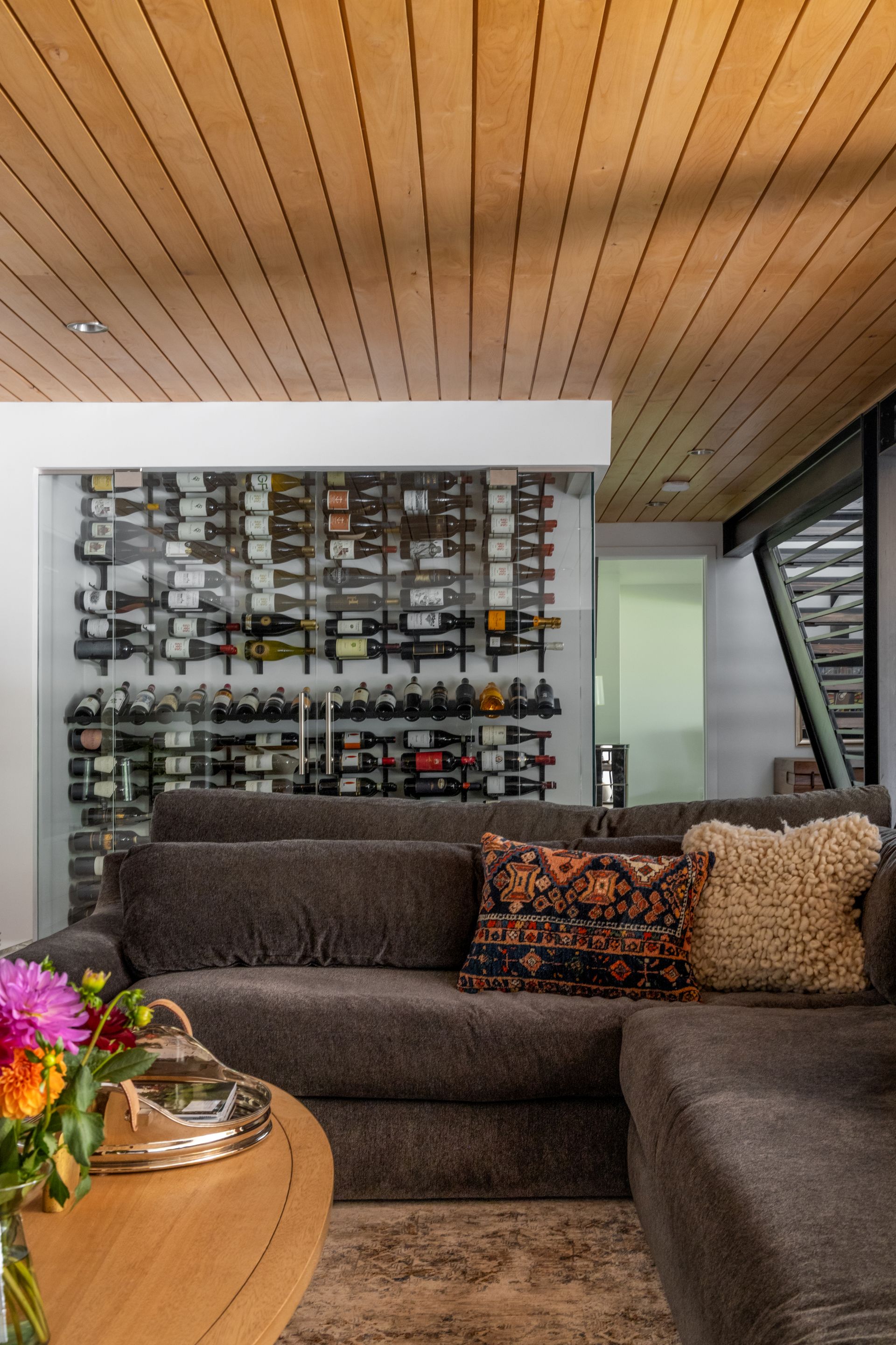 Living room with a glass-walled wine cellar, brown couch, and a wooden ceiling.