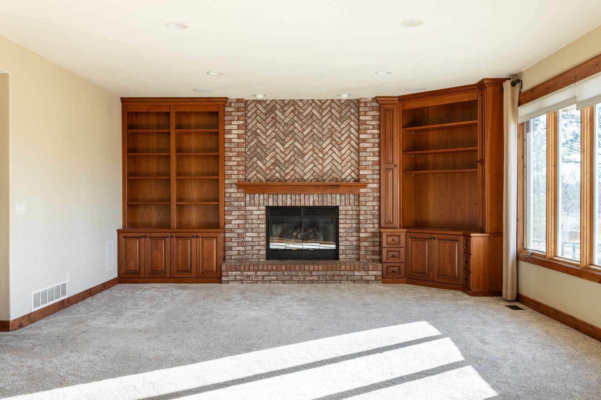 Empty living room with brick fireplace, built-in wood shelves, and a bay window.