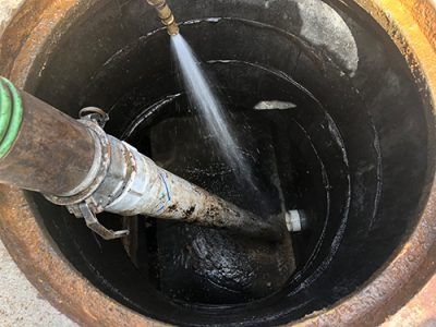 Jet of water cleaning the inside of a dark, circular manhole. A hose and nozzle are visible.