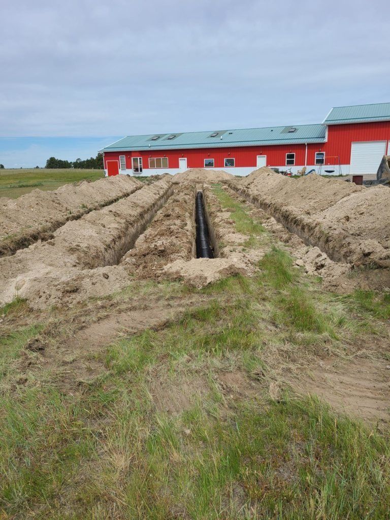 Trenches dug in a field in front of a red building, with pipes visible in one.