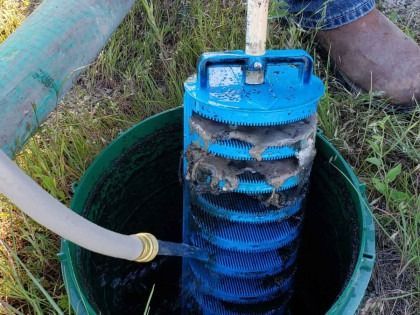 A blue well filter covered in mud being cleaned with a hose in a green bucket, outdoors.