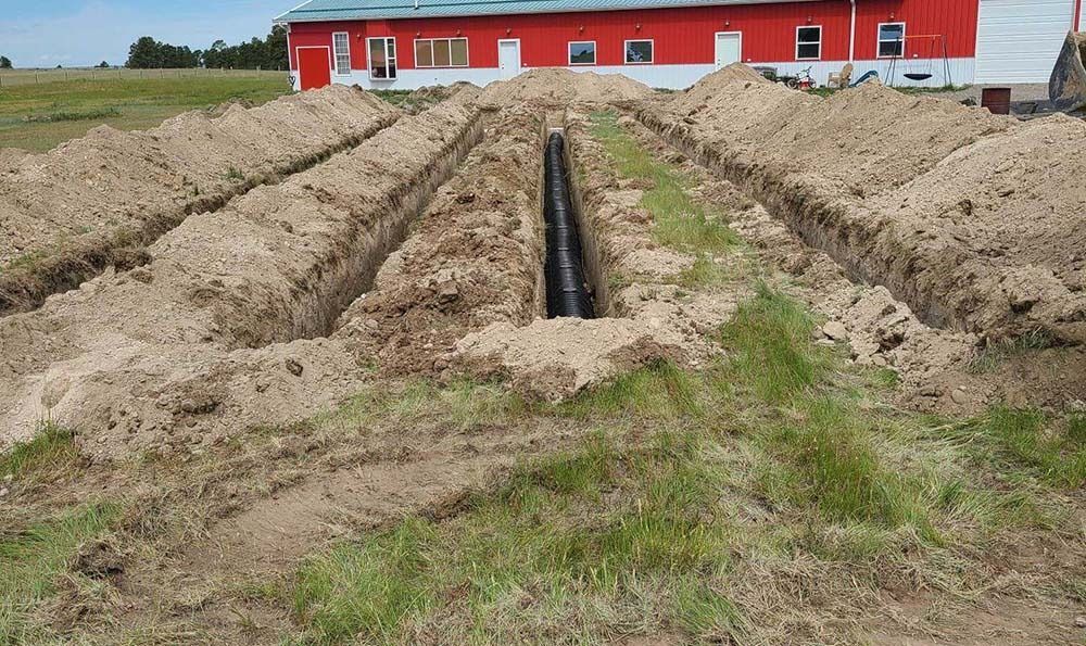 Trenches dug in a field with black pipe installed. A red building is in the background.
