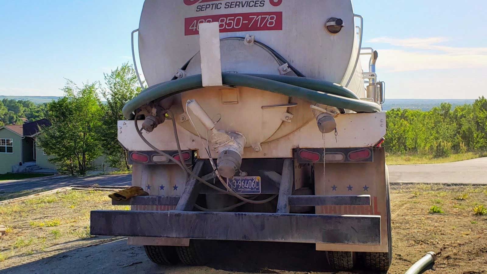 Rear view of a white septic tank truck with green hoses, parked on a gravel driveway, sunny day.