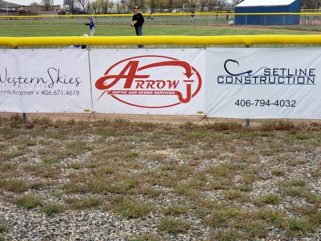 Three sponsor signs at a baseball field: Western Skies, Arrow J, and Setline Construction, with players in the background.