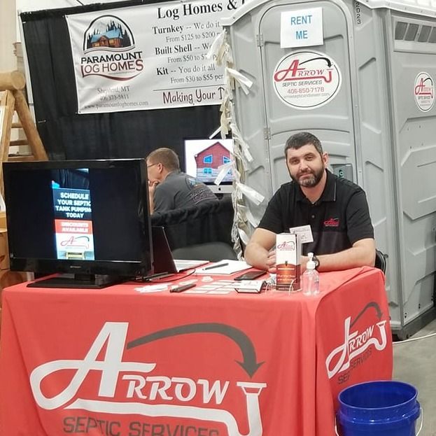 Man at Arrow Septic Services booth, red tablecloth, portable toilet in background.